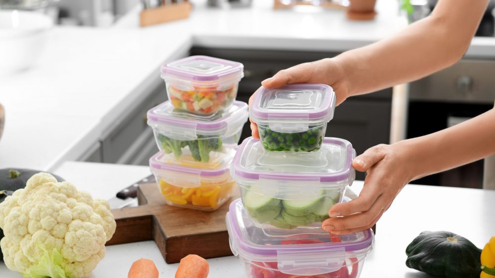 Woman holding stack of plastic containers with fresh vegetables for freezing at table in kitchen